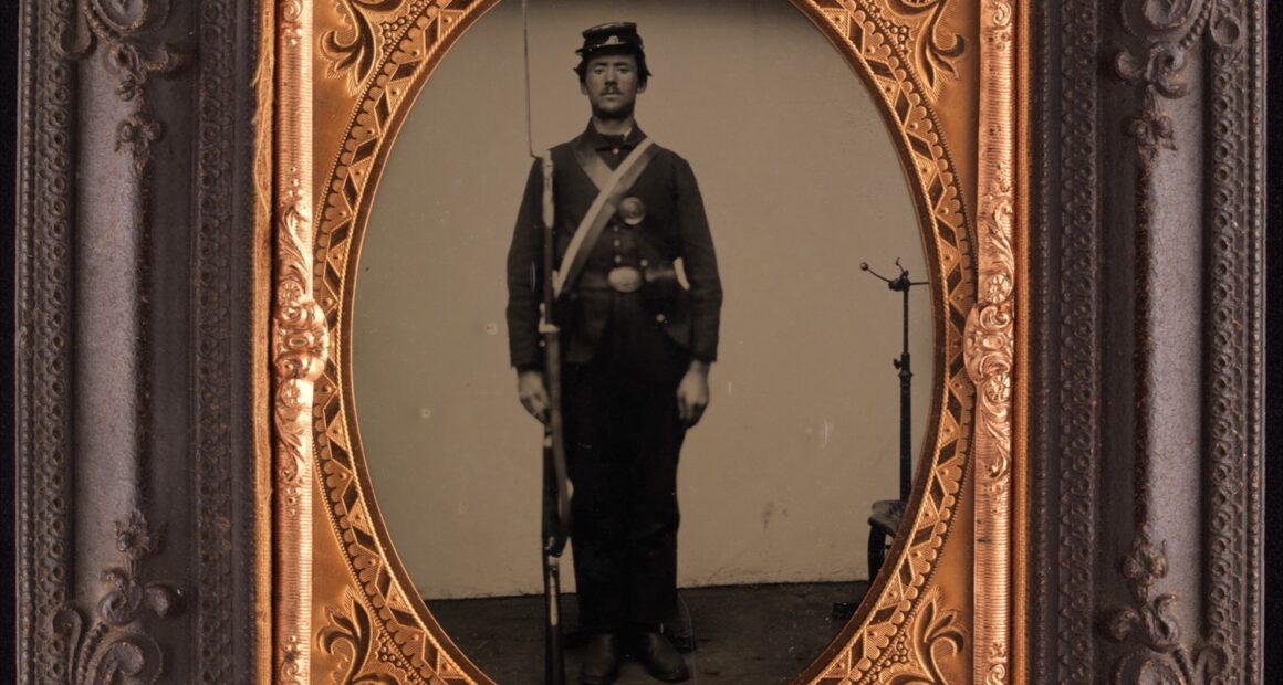 Tintype portrait of a standing Union soldier holding a rifle; a metal posing stand is visible behind him on the right.