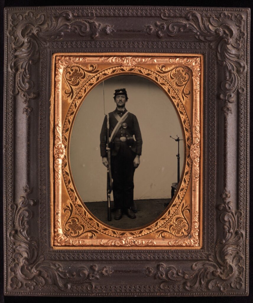 Tintype portrait of a standing Union soldier holding a rifle; a metal posing stand is visible behind him on the right.